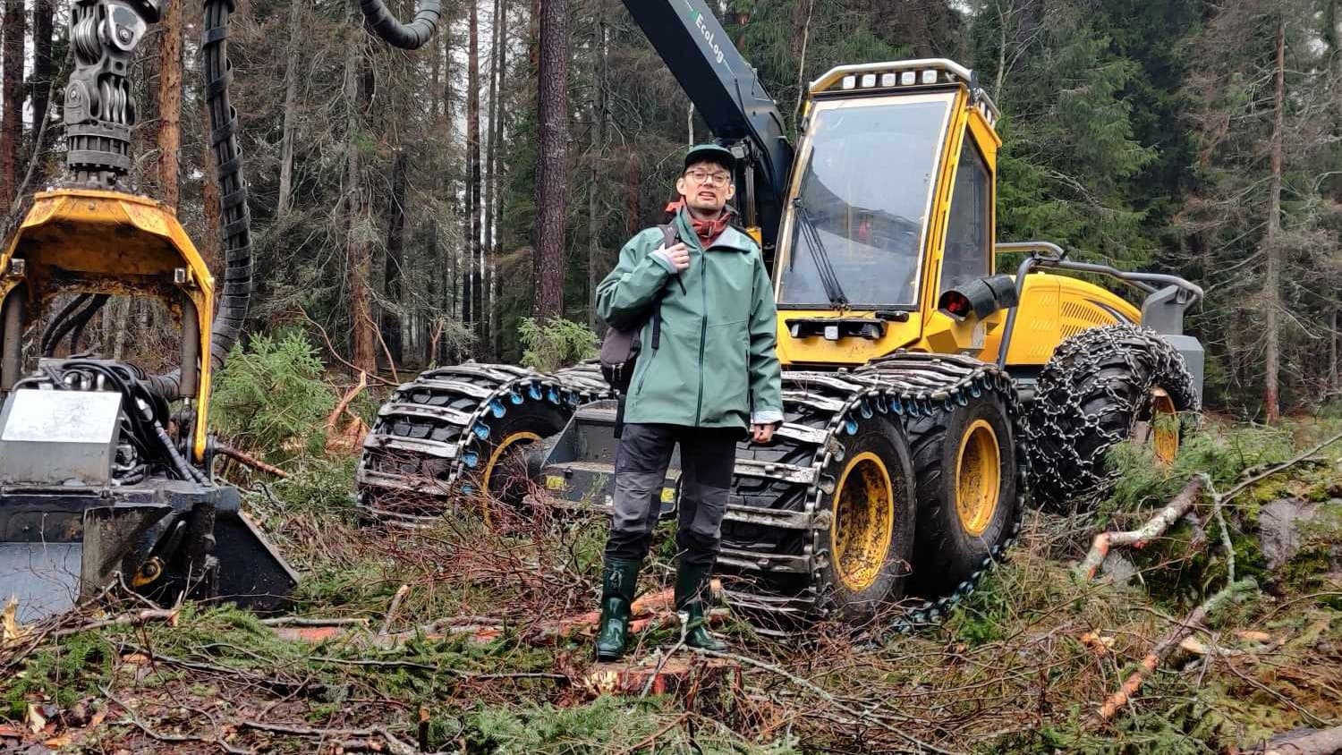 Person i grön jacka och gröna stövlar står framför en gul skogsmaskin i en skog med avverkade träd.