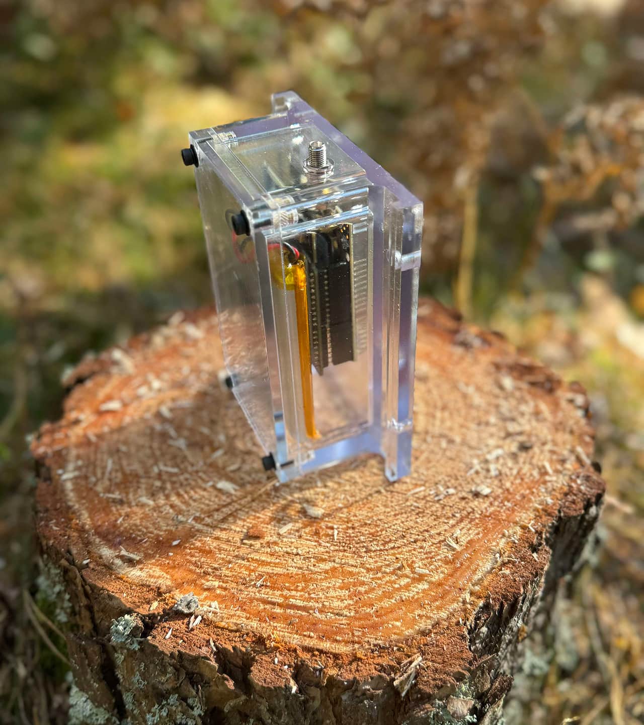 Elektronisk enhet i ett genomskinligt hölje placerad på en trädstubbe utomhus.