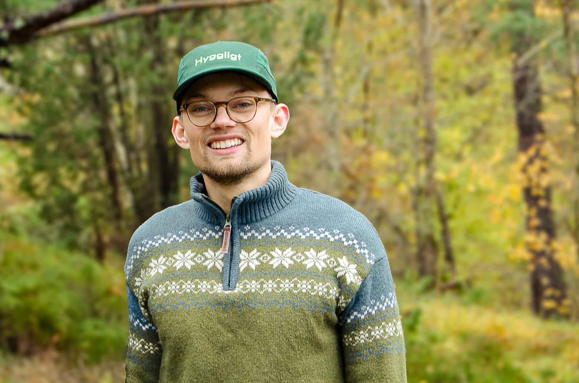 Man med keps med texten "Hyggligt" och en stickad tröja med mönster i skogsmiljö.